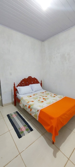 Ein Einzelbett mit floralem Bettbezug und einer orangefarbenen Decke, ein Holz-Kopfteil und ein Streifenmuster-Teppich auf Fliesenboden.