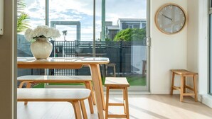 Una stanza moderna con un tavolo e sedie in legno, un vaso con fiori bianchi, una sveglia rotonda e vista sulla città attraverso una porta vetrata.