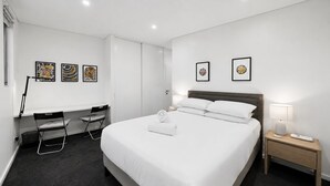 A hotel room with a large bed, a desk with a chair, two framed pictures on the wall, and a nightstand with a lamp and remote control. - Designer 1-Bed Apartment with Balcony (Rozelle)