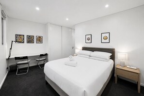 A hotel room with a large bed, a desk with a chair, a nightstand with a lamp, and two framed pictures on the wall.