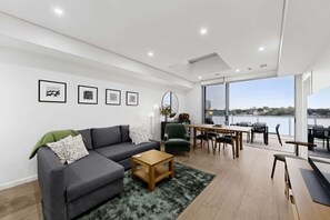 A modern living room with a sofa, coffee table, and dining area with chairs. There are framed pictures on the wall and a view of a body of water through large windows.