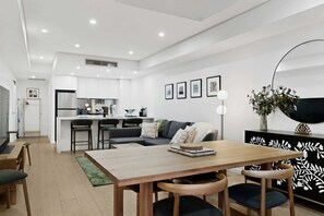 A modern kitchen with white cabinets, a dining area with a wooden table and chairs, and a living room with a sofa and a coffee table. - Designer 1-Bed Apartment with Balcony (Rozelle)