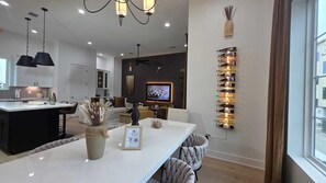 Cozinha moderna com ilha central, área de jantar com mesa branca e sala de estar com televisão e decoração na parede.