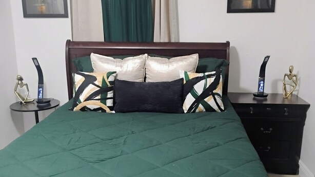 A bed with a green bedspread, two bedside tables with electronic devices, a wooden headboard, a dark-colored dresser, and two framed pictures on the walls. - WALKING DISTANCE FROM HARD ROCK STADIUM
1-bedroom apartment Miami Gardens (Miami Gardens)