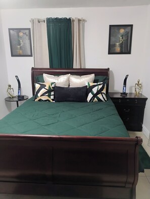 Una cama con colcha verde, dos mesitas de noche con dispositivos electrónicos, un cabecero de madera, un aparador de color oscuro y dos cuadros enmarcados en las paredes.