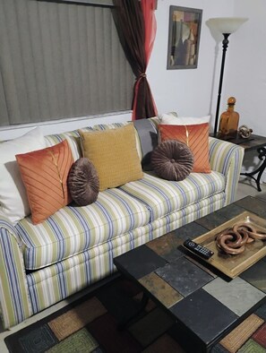 Ein gestreiftes Sofa mit mehreren Kissen, einer Fernbedienung und einem Tablett mit einer aufgerollten Kordel auf einem gefliesten Couchtisch.