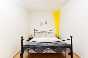 A bed with a metal frame and a patterned bedspread, a bedside table with a lamp, and a wall with two framed pictures.