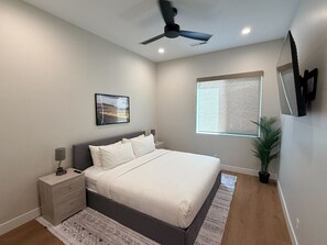 Ein Schlafzimmer mit einem Bett, Nachttischen, einem Deckenventilator, einem Fenster mit Jalousien, einer Topfpflanze und einem an der Wand montierten Fernseher.