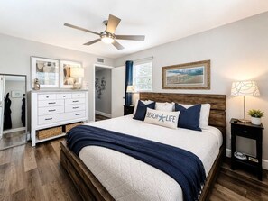 Ein Schlafzimmer mit Bett, Deckenventilator, Schminktisch, Nachttisch und einem gerahmten Bild an der Wand.