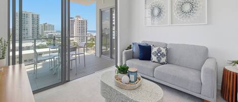 A modern living room with a grey sofa, a round coffee table with decorative items, and a view of the city through large glass doors.