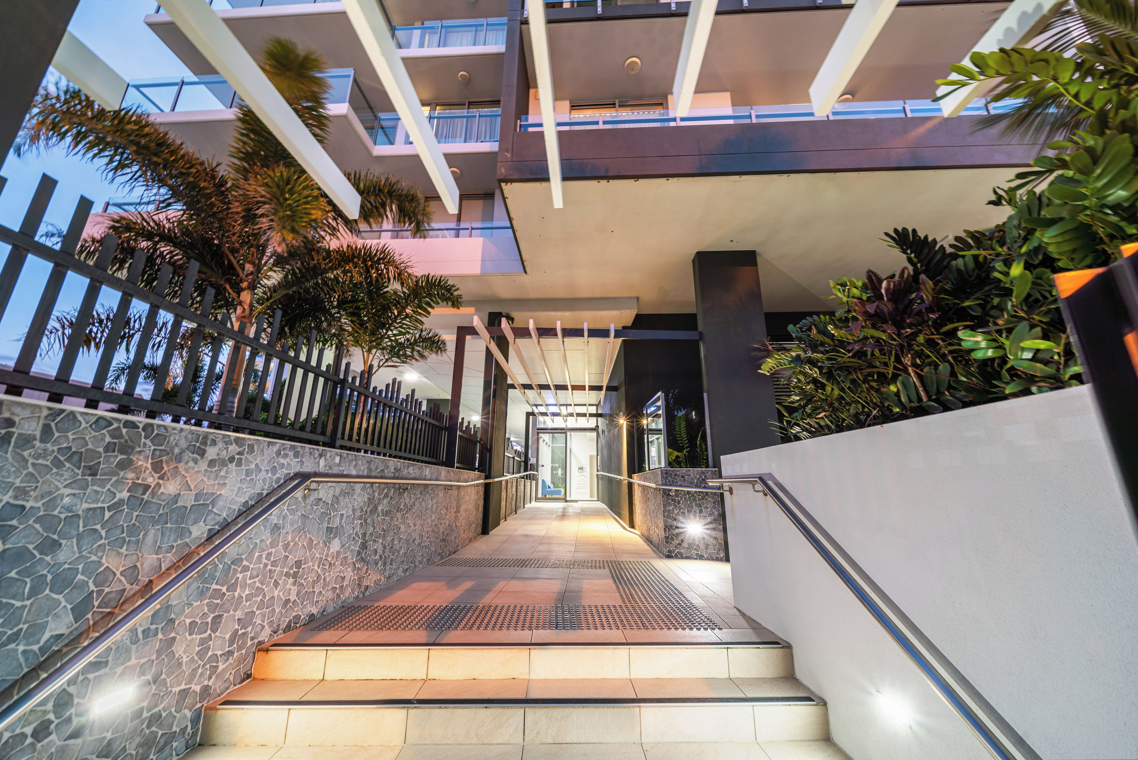A modern building entrance with a staircase leading to a covered area, surrounded by greenery and a stone wall.