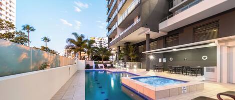 A modern hotel pool area with a spa, lounge chairs, and a view of surrounding buildings.
