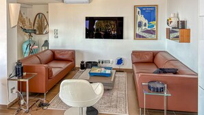 Uma sala de estar com dois sofás de couro, uma poltrona branca, uma mesa de centro, uma televisão e um quadro.