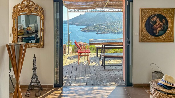 Uma porta aberta dá para uma varanda com vista para uma cidade costeira, chão de madeira, uma cadeira vermelha, uma mesa de madeira, um espelho decorativo, um quadro emoldurado e uma cesta de vime.