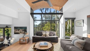 A modern living room with a large window, a ceiling fan, and a view of trees. - Pittwater Waterfront Retreat - Up to 10 Guests! (Coasters Retreat)
