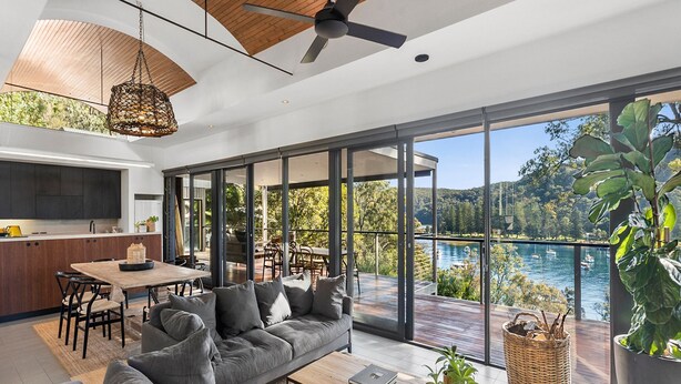 A modern interior with a dining area, sofa, and large windows overlooking a lake. - Pittwater Waterfront Retreat - Up to 10 Guests! (Coasters Retreat)