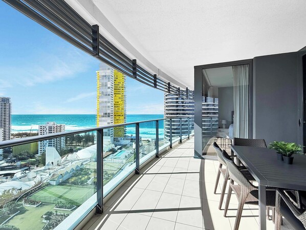 Een balkon met uitzicht op zee en stadsbeeld, een eethoek met buitentafels, en een modern gebouw met een glazen gevel.