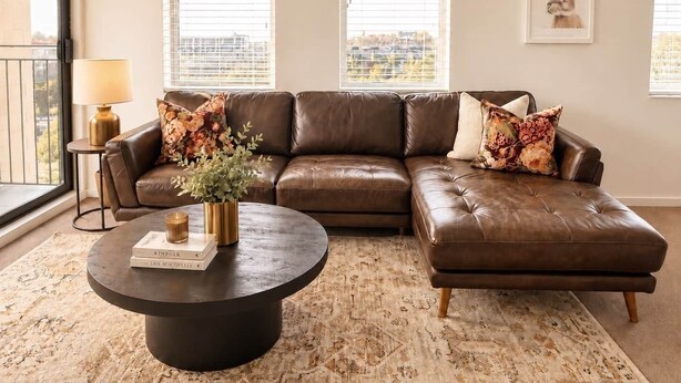 A brown leather sectional sofa with floral cushions, a round coffee table with books, and a city view through large windows. - Central CBD | Walk to Te Pae, Stadium + Parking (Christchurch)