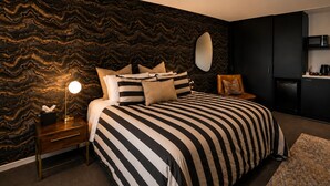 A bedroom with a striped bedspread, a bedside lamp, a dresser, a mirror, and a kitchen area with a microwave.