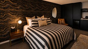 Ein Schlafzimmer mit gestreifter Bettdecke, einer Nachttischlampe, einem Schminktisch und einer Küchenzeile mit einer Mikrowelle.