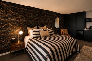 Een slaapkamer met een gestreept beddegoed, een bedlamp, een dressoir, een spiegel en een keukenhoek met een magnetron.