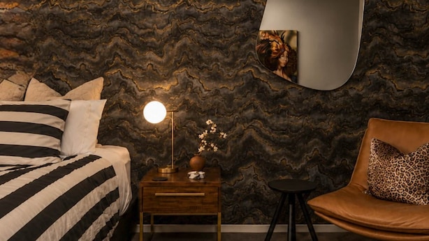 Ein Schlafzimmer mit Bett, Nachttisch, Lampe, Blumen in einer Vase, Stuhl und einer Wand mit einem gewellten Muster.