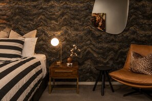Une chambre à coucher comprenant un lit, une table de chevet, une lampe, un vase avec des fleurs, une chaise et un mur orné d’un motif ondulé.