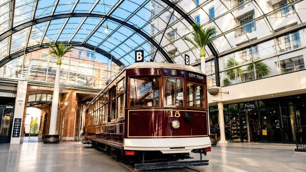 A vintage tram labeled 'CITY TOUR' with the number 15, parked inside a modern building with a glass roof and palm trees. - Central Apt | Walk Te Pae + Parking (Christchurch)
