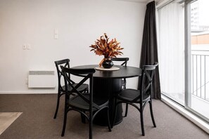 Ein runder Tisch mit vier schwarzen Stühlen, eine Vase mit Trockenpflanzen, ein weißer Heizkörper und ein großes Fenster mit Blick auf Gebäude.