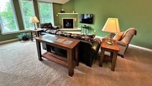 Una sala de estar con sofá, mesa de centro, chimenea, televisor y ventana con vistas a árboles.