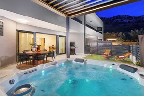 A modern outdoor hot tub area with a dining table and chairs, surrounded by a patio with lounge chairs. - Luxury suite Ema with jacuzzi (Draga Bašćanska)