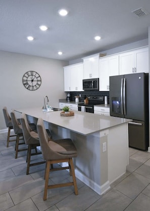 A modern kitchen with white cabinets, a stainless steel refrigerator, a microwave, and a bar area with stools. - Luxury Disney Retreat: Pvt Pool + Resort Access (Kissimmee)