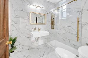 Un baño con paredes de mármol, un lavabo blanco, un espejo con marco dorado, una ducha con cerramiento de vidrio y un inodoro.