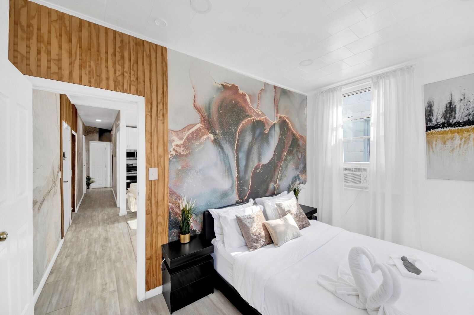 A hotel room with a bed, a large abstract painting, a wooden panel wall, and a view of the outside.