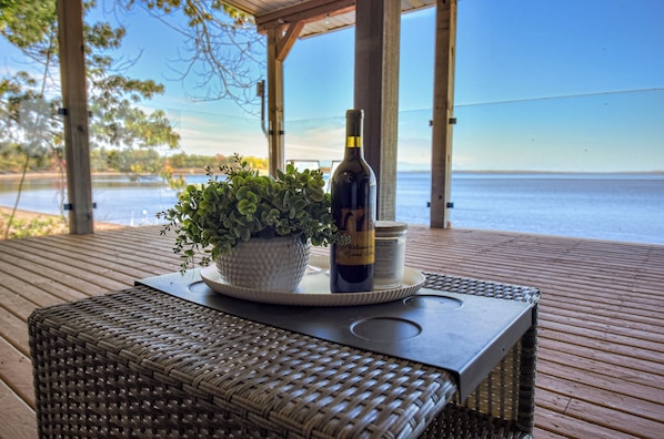 A wicker table with a bottle and a potted plant on a wooden deck overlooking a body of water.