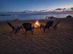 Escena de playa con fuego, sillas y un barco al fondo.
