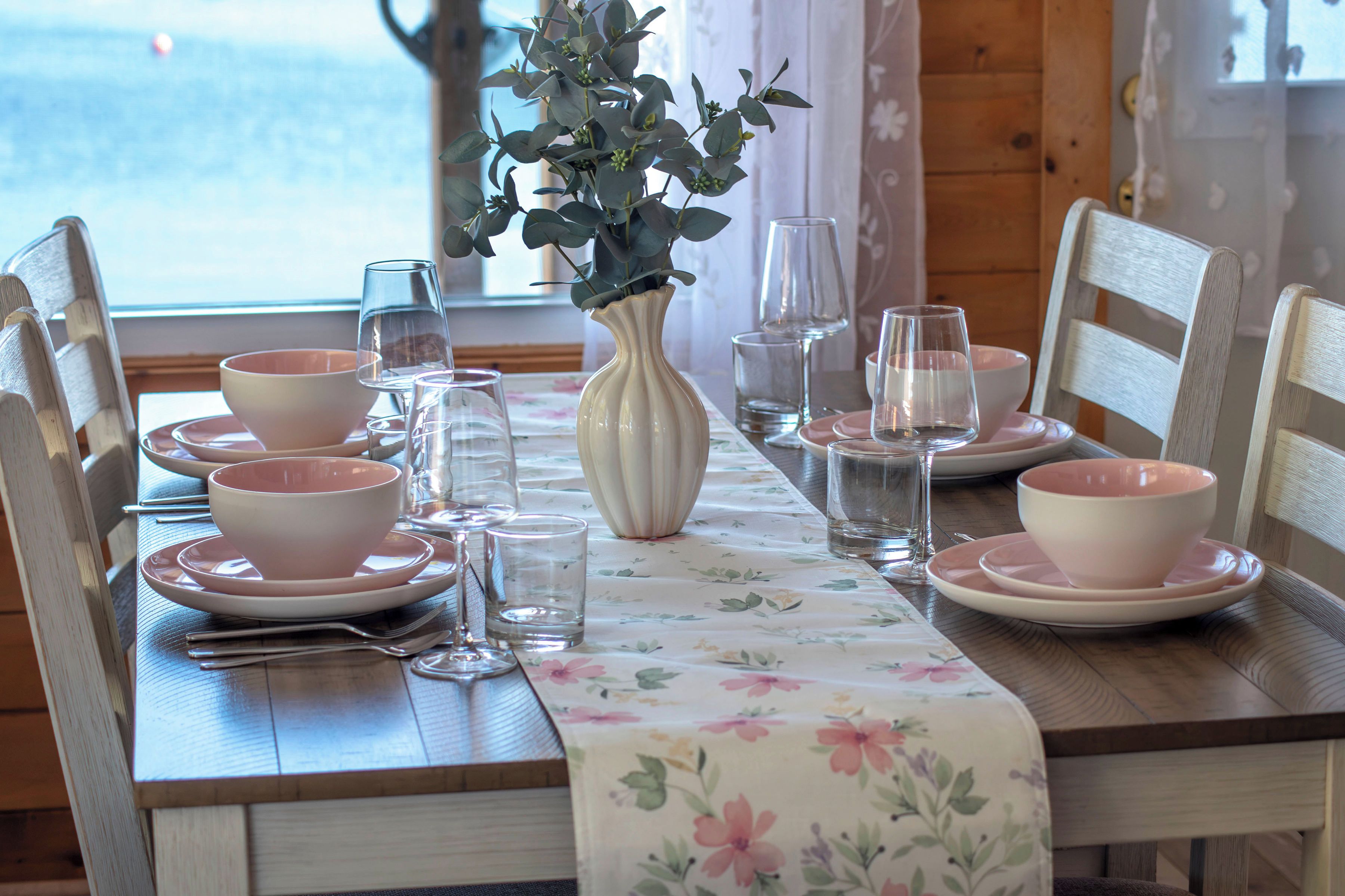 Une table à manger dressée avec des bols, des assiettes et des verres transparents de couleur rose, et un vase rempli de feuillage vert placé au centre.