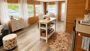A modern kitchen with wooden cabinets, a dining area with a table and chairs, a small kitchenette, and a view of greenery outside. - Cozy Beach Escape/Fire pit/Sunrises/Lake Views/Golf/Pickleball/45 to Fredericton (Canning)