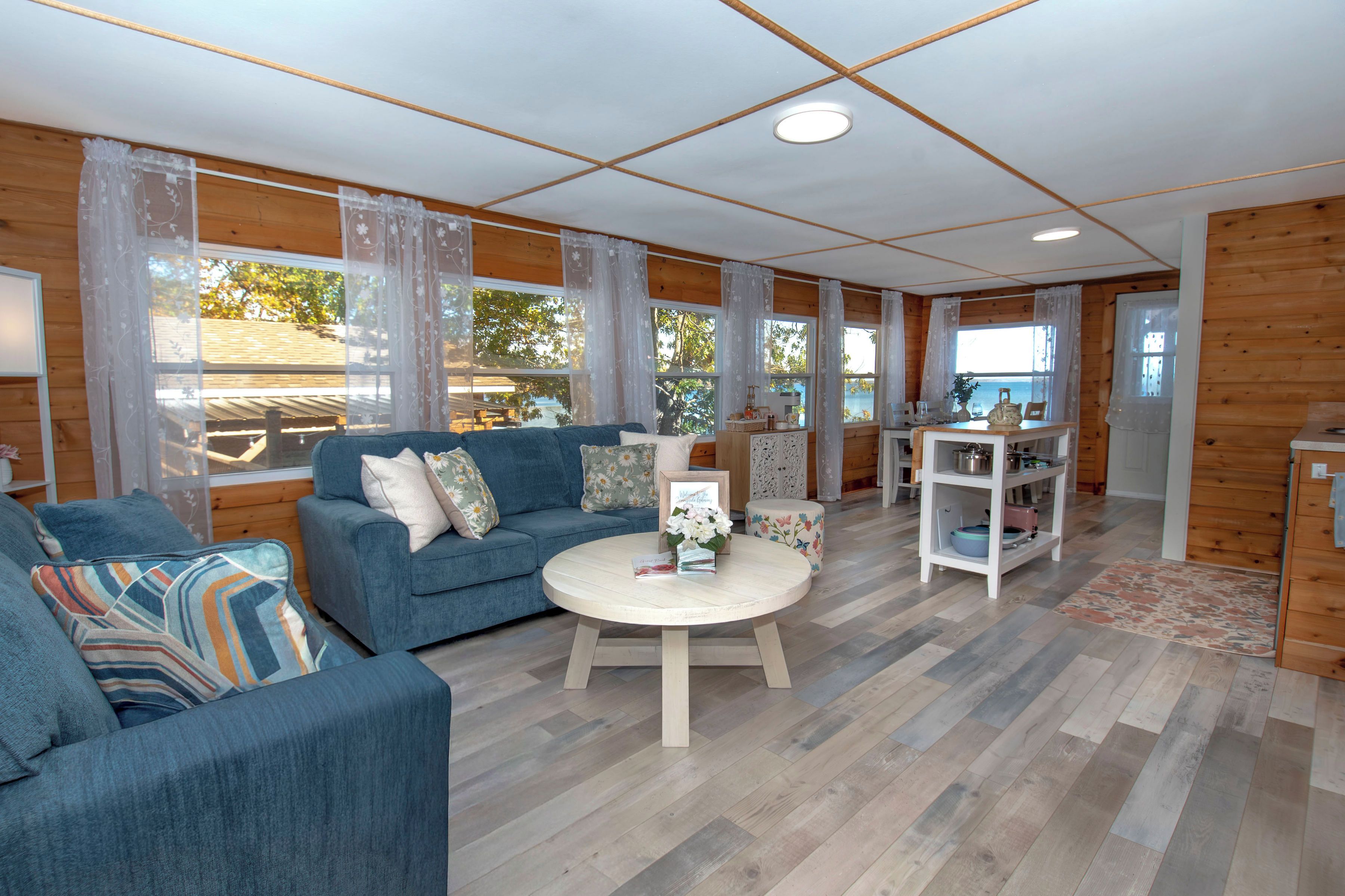 A cozy living room with a blue sofa, a round coffee table, and a kitchen area with wooden cabinets and a white shelf.