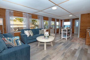 A cozy living room with a blue sofa, a round coffee table, and a kitchen area with wooden cabinets and a white shelf.