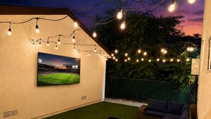 Um pátio externo com grama sintética, uma televisão montada na parede, móveis de vime e luzes de festa.