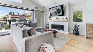 A modern living room with a grey sofa, a flat-screen TV, a fireplace, and a balcony with a view of buildings and trees. - South Bay Sweet Spot: Walk to Hermosa & MB Piers 5 bdrm home 2 Blocks from Beach (Hermosa Beach)