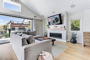 Un soggiorno moderno con un divano grigio, una TV a schermo piatto, un camino e un balcone con vista su edifici e alberi.