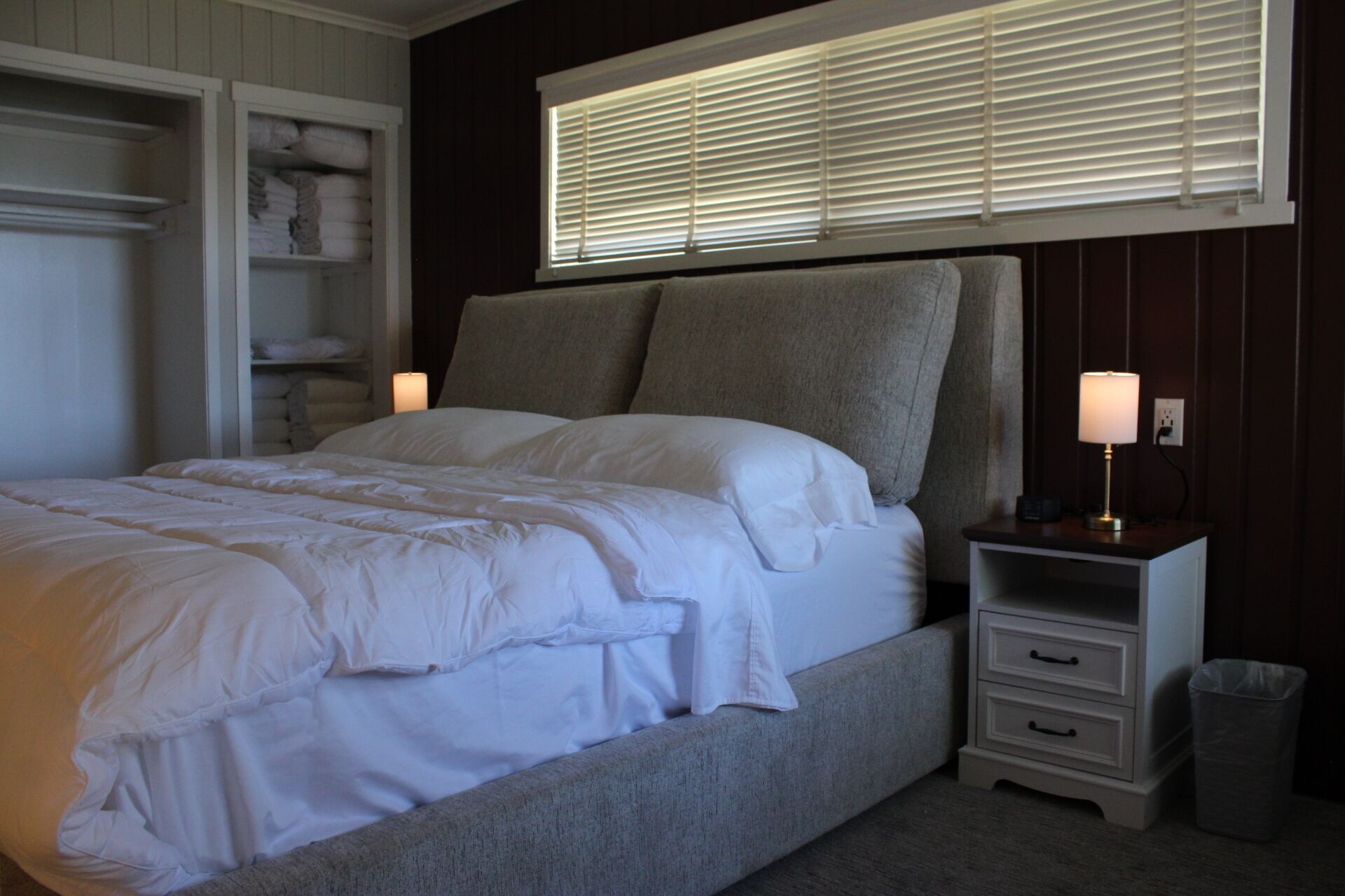 Une chambre à coucher comprenant un lit, une table de chevet, une lampe, une fenêtre avec des stores et une armoire.