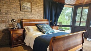 A bedroom with a wooden bed, a nightstand, a dresser, a window with curtains, and a brick wall. - Apple Tree Studio - country luxury self-contained studio (Kilmore)