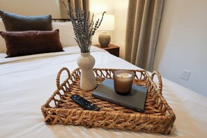 Ein ordentlich bezogenes Bett mit Fernbedienung, einem Buch und einer Kerze in einem gewebten Korb.
