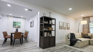 Un salon moderne avec une bibliothèque en bois foncé, un canapé beige, un coin repas avec une table et des chaises, et une grande fenêtre avec des rideaux.