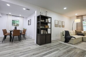 Ein modernes Wohnzimmer mit einem dunklen Holzregal, einem beigefarbenen Sofa, einem Essbereich mit Tisch und Stühlen sowie einem großen Fenster mit Vorhängen.