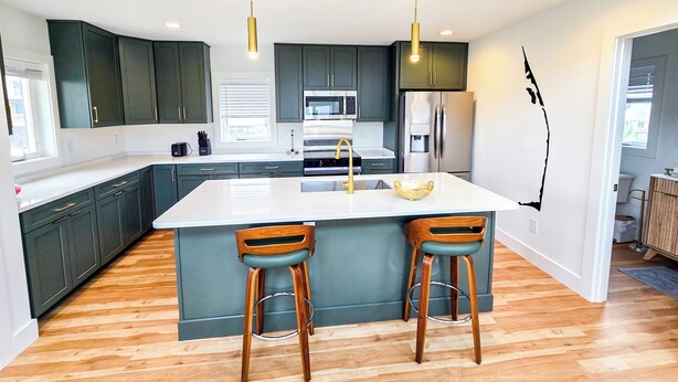 A modern kitchen with dark green cabinets, a white island, two wooden bar stools, and stainless steel appliances. - Cozy 1 bedroom house in serene Kitty Hawk with Rooftop views of Ocean and sunset (Kitty Hawk)