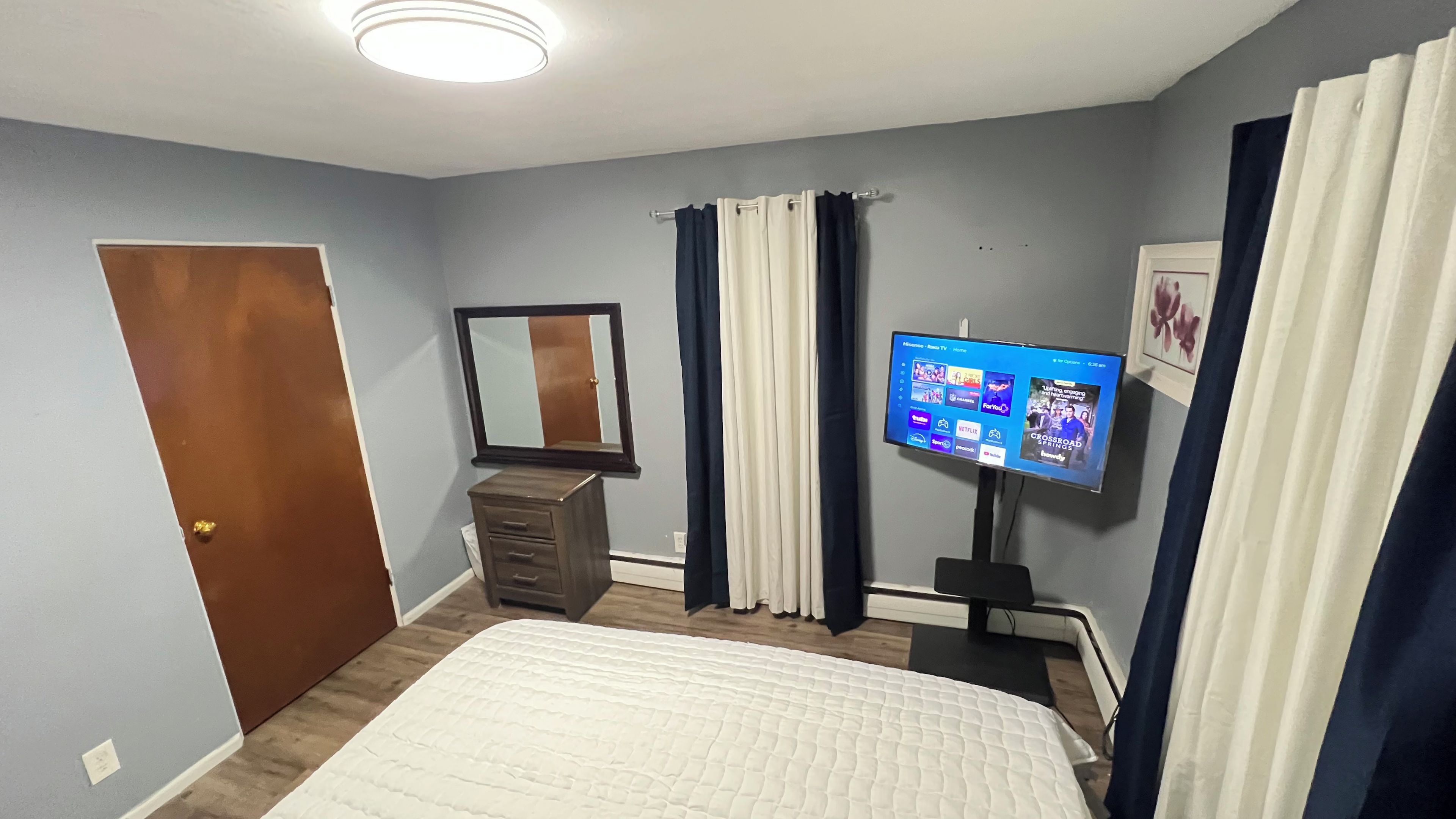 A bedroom with a bed, dresser, mirror, curtains, and a TV displaying a home screen.